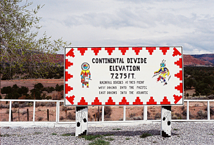 [A large white sign supported by a post on either end is several feet above the ground. There are red mesa-like symbols around the border of the sign and a Native American dancer symbol at each end. The sign says, 'Continental Divide Elevation 7,275 feet. Rainfall divides at this point. West drains into the Pacific. East drains into the Atlantic.' There are outlines of feather symbols on the posts.]